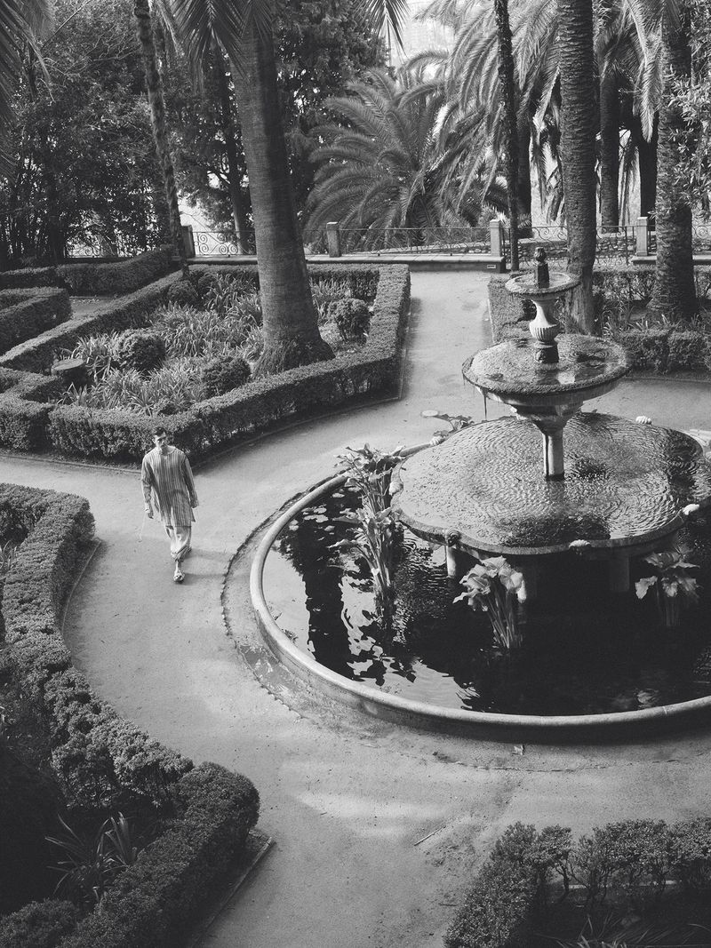 High-angle black and white view of model Xavier Serrano walking along a garden path beside a tiered stone water fountain.