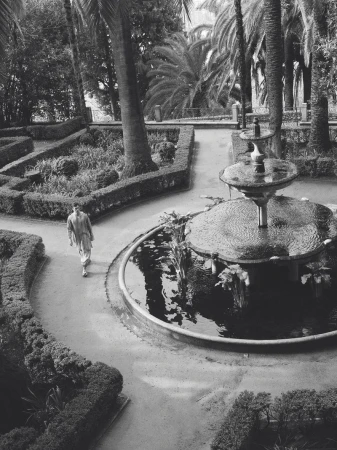 High-angle black and white view of model Xavier Serrano walking along a garden path beside a tiered stone water fountain.