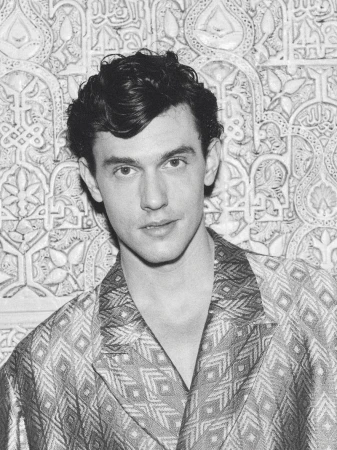 Monochrome close-up portrait of model Xavier Serrano wearing a geometric patterned jacket against an ornate carved relief wall.