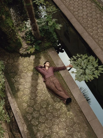 Model Xavier Serrano lying beside a reflective garden water canal wearing a plum tunic shirt and textured brown wide-leg trousers.