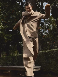 Xavier Serrano wearing a draped beige suit while balancing on a stone garden bench among citrus trees.
