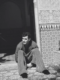 Model Xavier Serrano seated on the ground beside ornate mosaic tilework in a black and white editorial portrait.