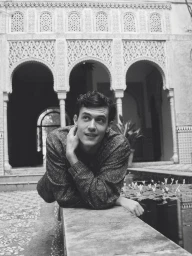 Model Xavier Serrano leaning over a stone ledge in front of ornate Moorish arches in a black and white fashion portrait.