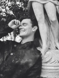 Black and white photo of model Xavier Serrano smiling and leaning against a classical stone statue outdoors.