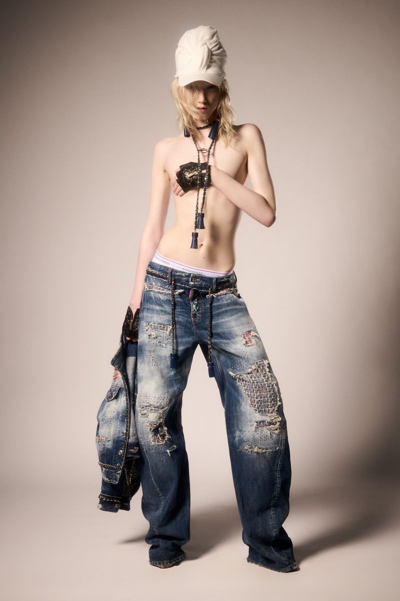 Fashion photo of a slender person with a white turban, wide-leg distressed jeans, bolo tie, and black gauntlets in a photo studio.