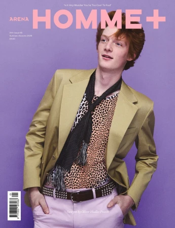 A red-haired young man in a beige blazer, leopard print top, black knit scarf, and pink trousers poses against a lilac background.
