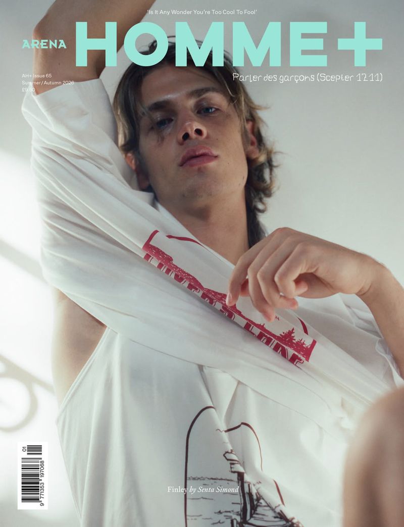 A young man lifting a white graphic t-shirt over his head for the cover of Arena Homme+ magazine against a neutral wall.