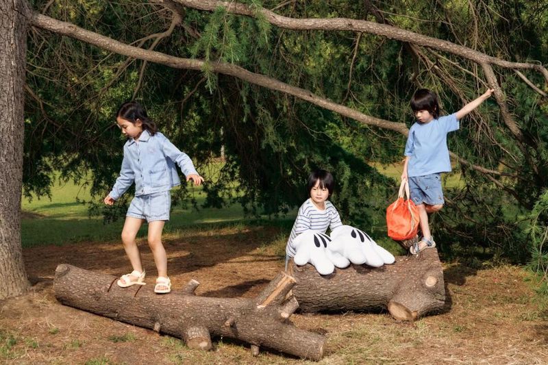 Three kids playing on a fallen log in a sun-dappled park. One girl balances on the log, a boy stands with a bag, and another sits.