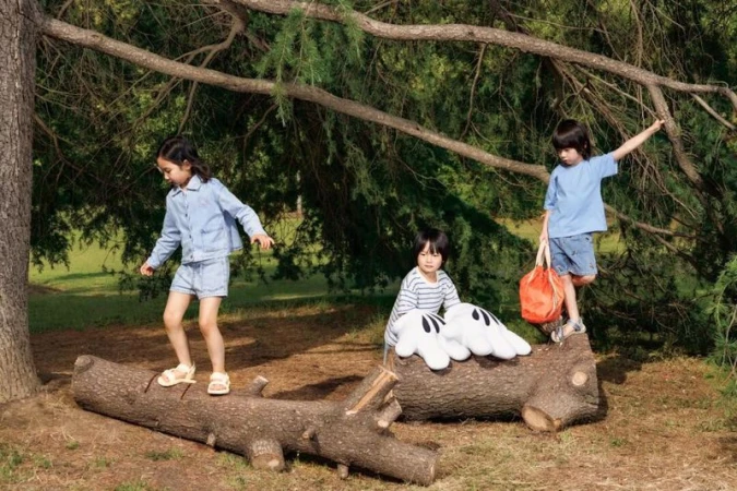 Three kids playing on a fallen log in a sun-dappled park. One girl balances on the log, a boy stands with a bag, and another sits.