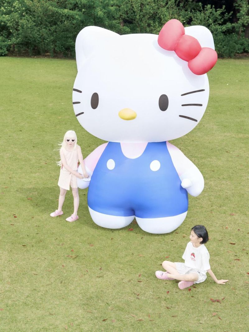Giant Hello Kitty inflatable with two young women in a park setting, one standing and the other sitting on green grass.