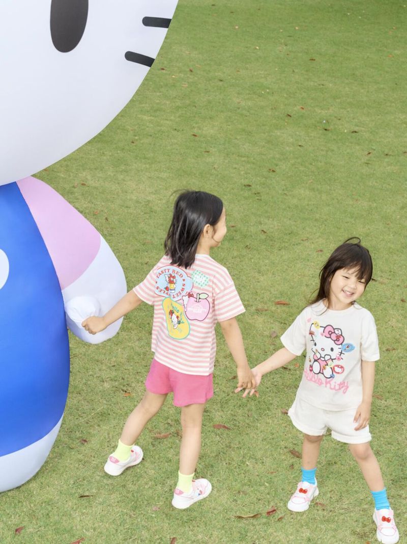 Two children wearing Hello Kitty apparel hold hands in a grassy outdoor setting near a large inflatable Hello Kitty figure.