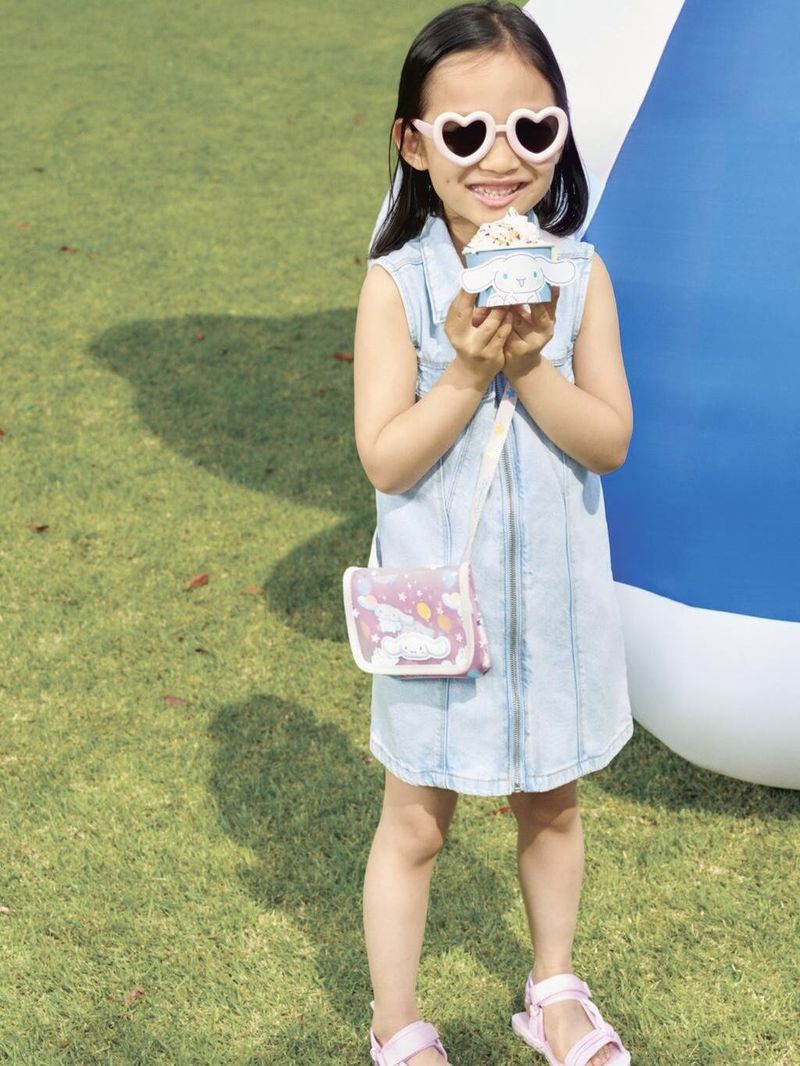 Young girl in a denim dress and heart-shaped sunglasses holds an ice cream treat in a park-like outdoor setting with green grass.