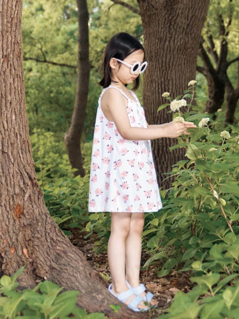 A young girl in a white patterned dress and sunglasses stands in a green forest, looking at white flowers, with trees in the background.