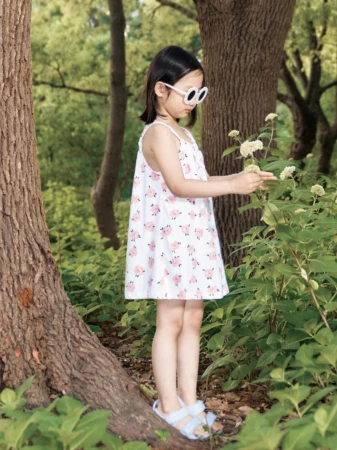 A young girl in a white patterned dress and sunglasses stands in a green forest, looking at white flowers, with trees in the background.