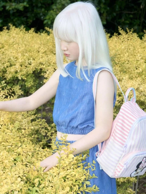 A young, fair-skinned child with white-blonde hair stands outside amidst yellow flowering bushes, wearing a blue denim outfit and pink-striped backpack.