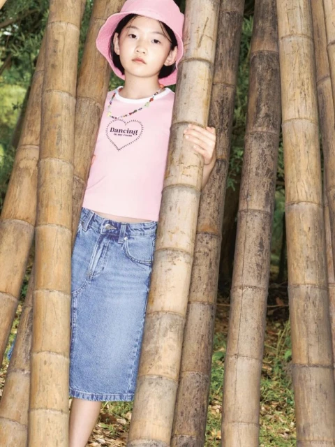 A young girl poses next to bamboo stalks, wearing a pink top, denim skirt, and a pink bucket hat in an outdoor setting.
