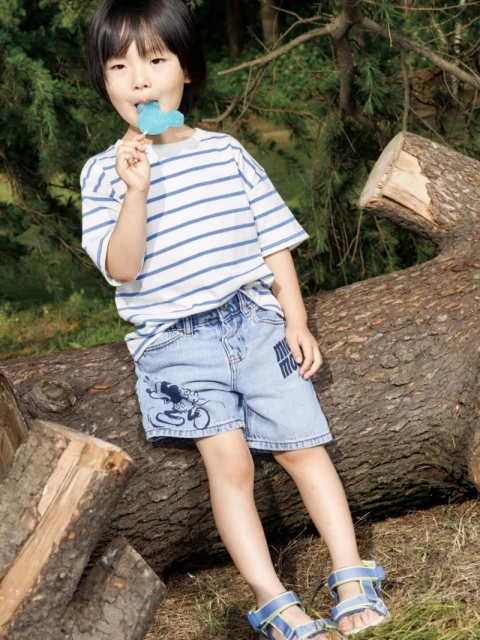 A young Asian child sits on a log outdoors eating a blue popsicle, wearing a striped shirt, denim shorts, and blue sandals.