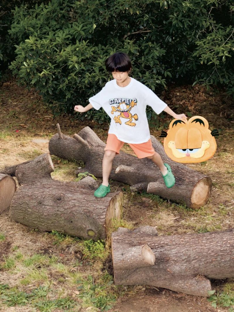 A young child balances on logs outdoors, wearing a Garfield T-shirt, shorts, and clogs, while holding a Garfield-themed cushion.