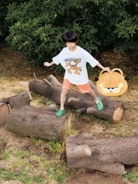 A young child balances on logs outdoors, wearing a Garfield T-shirt, shorts, and clogs, while holding a Garfield-themed cushion.