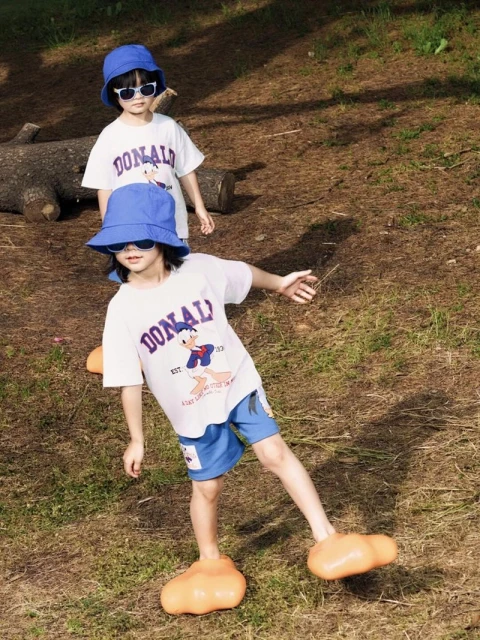 Two kids, a boy and a girl, standing on grass in a park wearing matching Donald Duck shirts, blue shorts, bucket hats, and sunglasses.