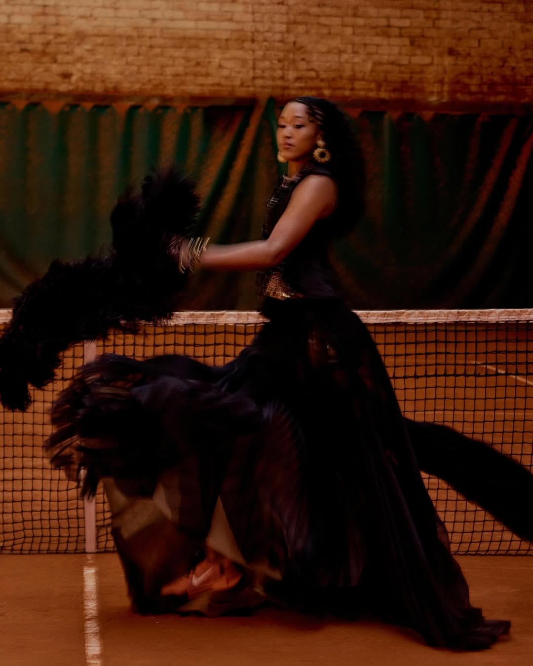 Naomi Osaka posing in an ornate black gown with dramatic feather accents beside a net on an indoor tennis court.