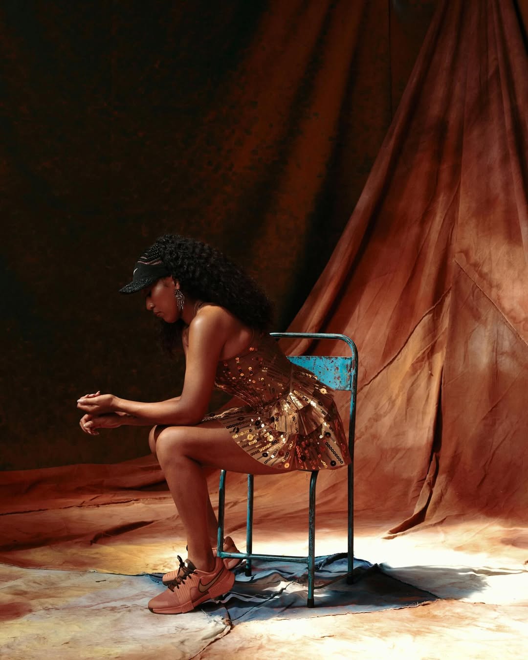 Naomi Osaka seated in profile on a turquoise chair wearing a gold sequin dress and black visor against a warm draped backdrop.