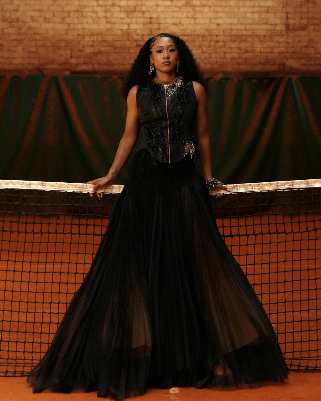 Naomi Osaka posing behind a tennis net wearing an embellished sleeveless black gown on a clay court.