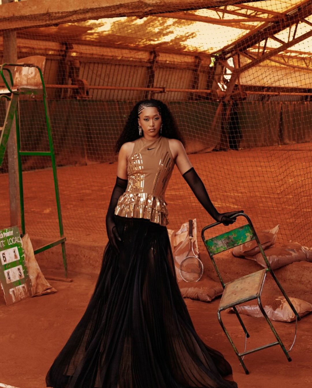 A woman in a gold sequin top and black skirt poses on a tennis court near a chair and a scoreboard, with an earthy, vintage aesthetic.