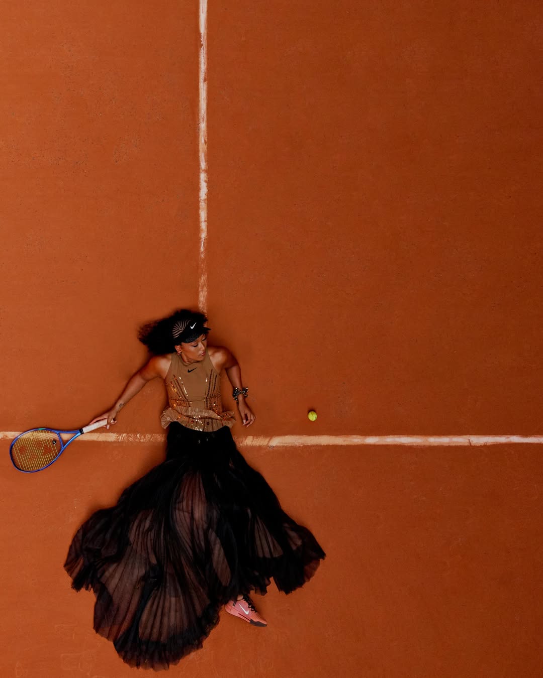 Naomi Osaka lying on an orange clay tennis court in a black tulle gown holding a tennis racket with a tennis ball nearby.