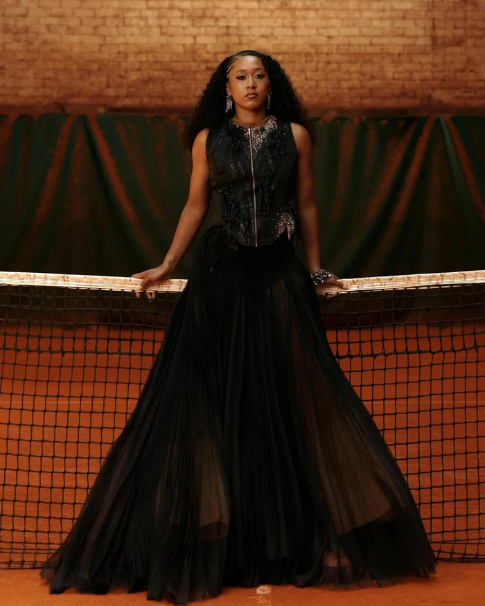 Naomi Osaka posing behind a tennis net wearing an embellished sleeveless black gown on a clay court.