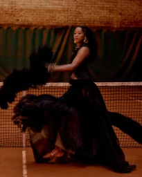 Naomi Osaka posing in an ornate black gown with dramatic feather accents beside a net on an indoor tennis court.