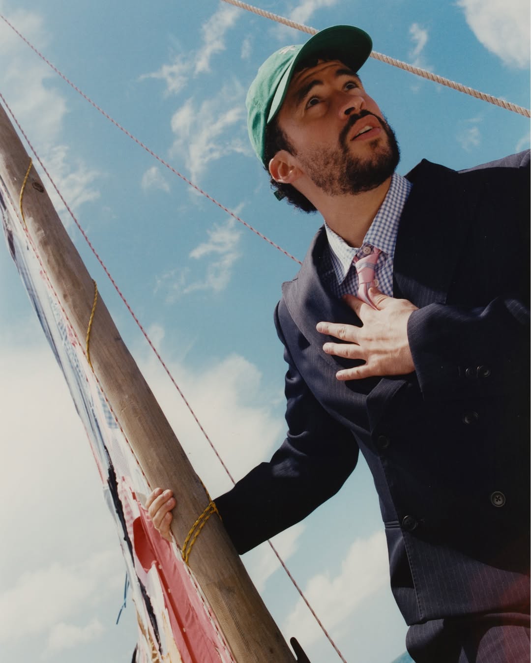 Bad Bunny wearing a green baseball cap and dark tailored suit holding a wooden sailboat mast against a blue sky.