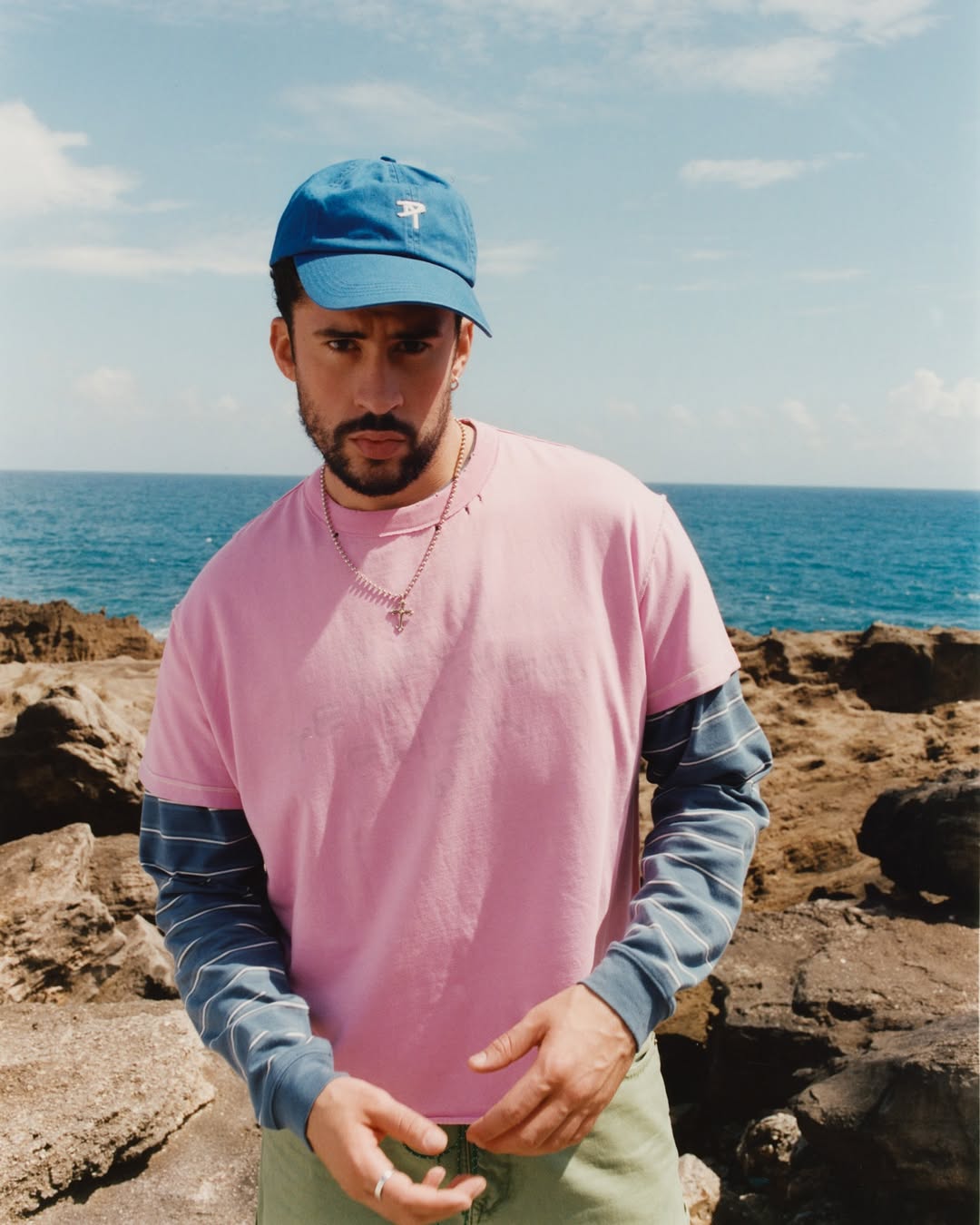 Bad Bunny posing on a rocky coastline wearing a pink t-shirt over striped sleeves, a blue cap, and a silver necklace.