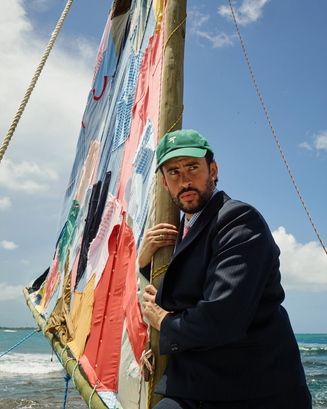 Bad Bunny wearing a navy blazer and green baseball cap holds the wooden mast of a sailboat with a patchwork shirt sail by the ocean.
