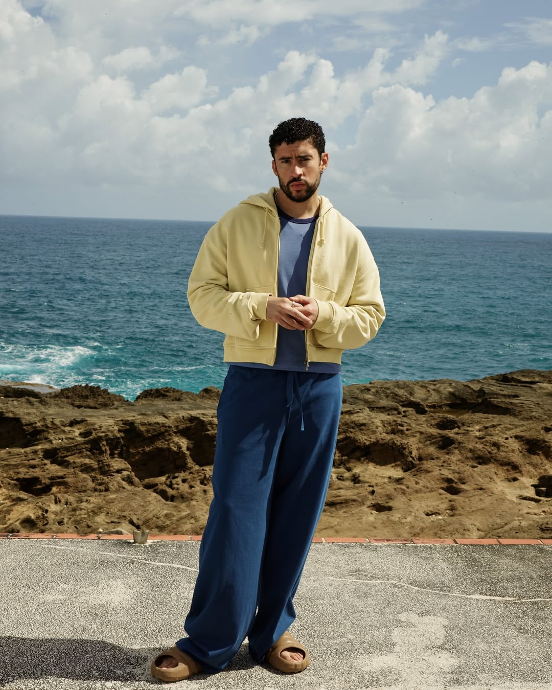 Bad Bunny wearing a yellow hoodie and blue trousers standing against an ocean and rocky coast backdrop for Zara