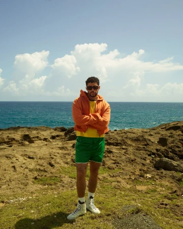 Bad Bunny stands confidently on a rocky coast with the ocean and sky in the background, wearing sunglasses and a colorful casual outfit.