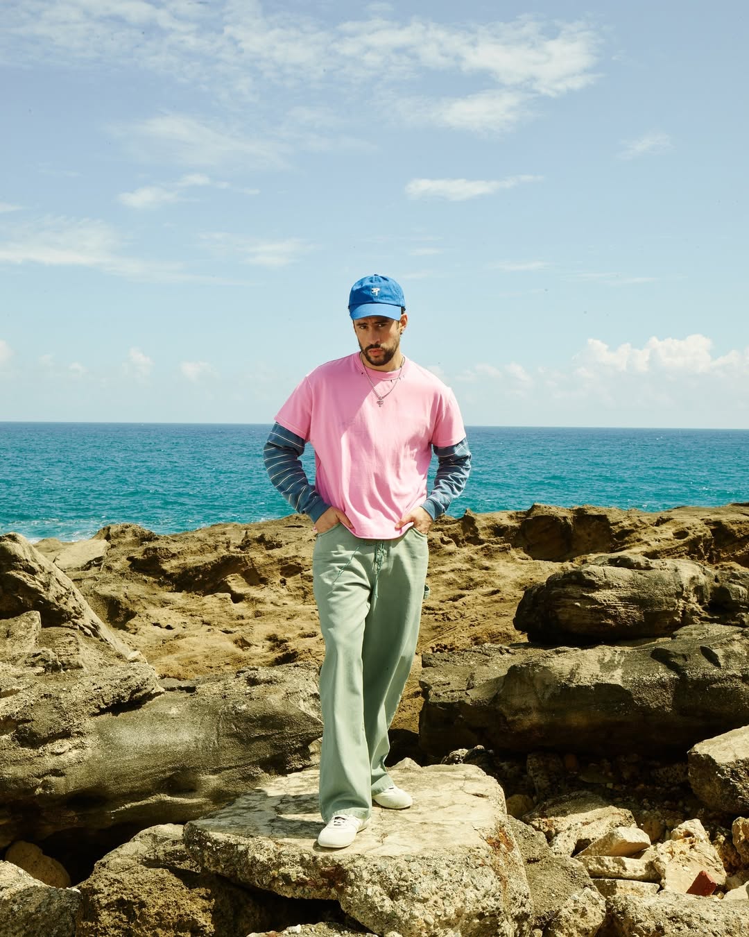 Bad Bunny stands on rocks near the ocean wearing a pink t-shirt, blue cap, and light green pants under a sunny, blue sky.