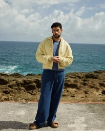 Bad Bunny wearing a yellow hoodie and blue trousers standing against an ocean and rocky coast backdrop for Zara