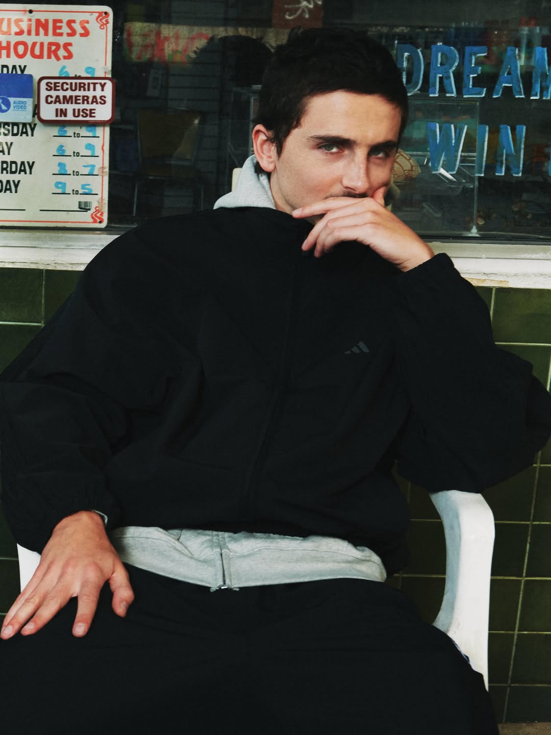 Young man seated in a white chair wearing a black jacket and grey hoodie outdoors against a storefront window.