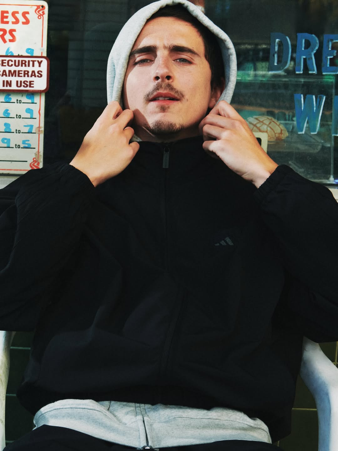 Man wearing a grey hood under a black Adidas zip-up jacket adjusting his hood while seated in front of a shopfront window.