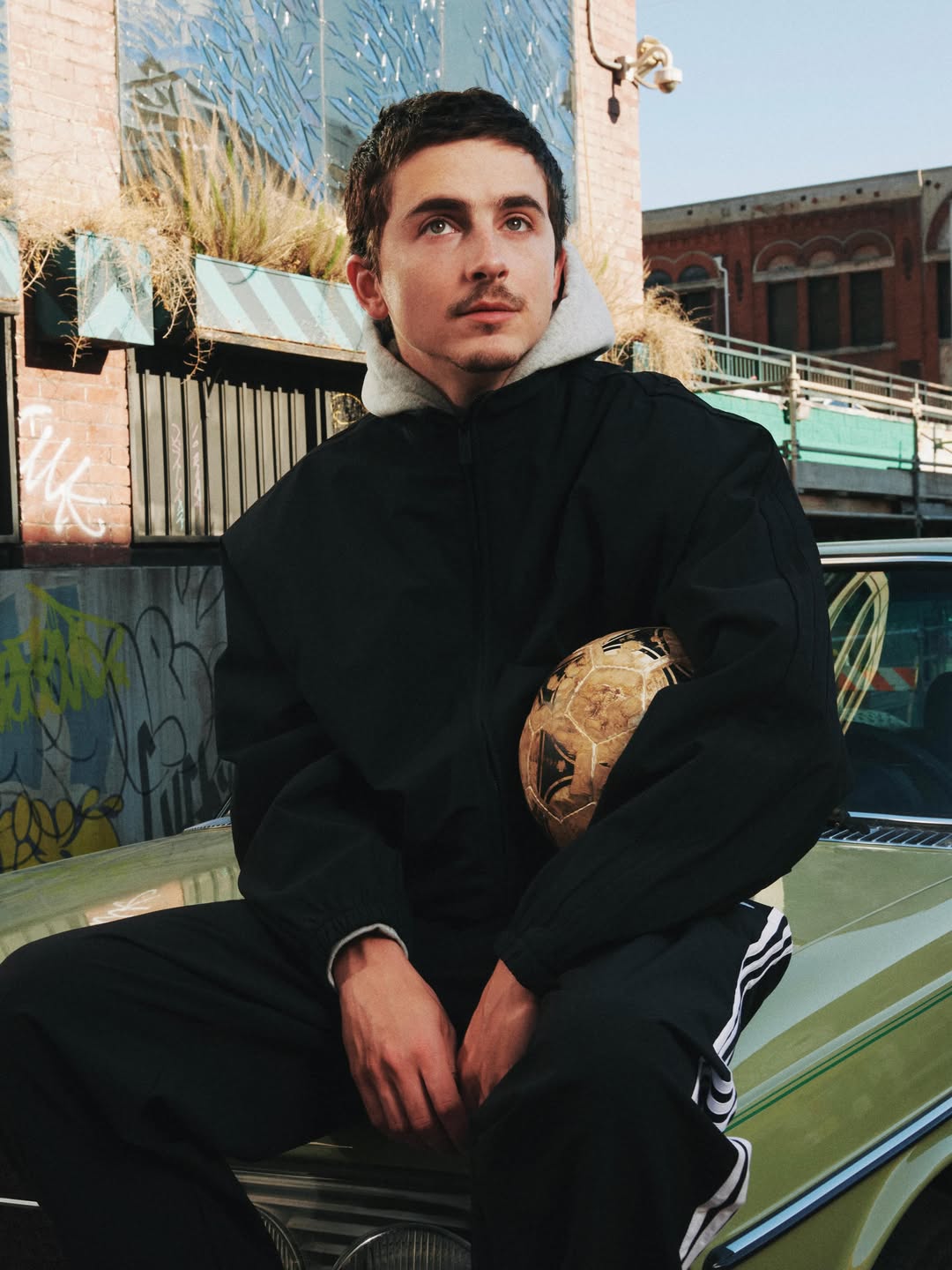Young man in a black jacket and Adidas track pants holding a soccer ball while sitting on the hood of a vintage car outdoors.
