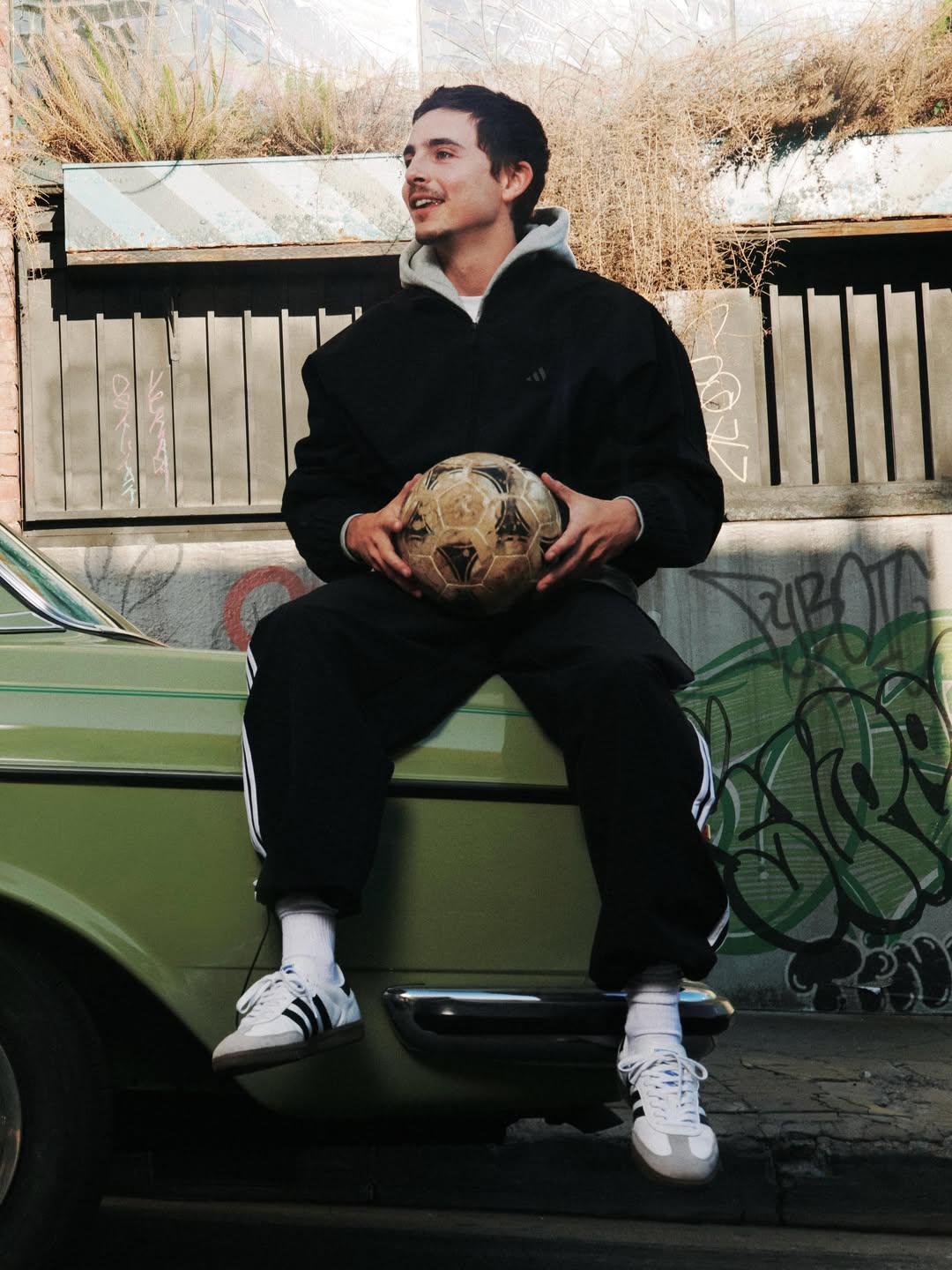 Young man in a black Adidas tracksuit and sneakers sits on the hood of an olive green vintage car holding a soccer ball.