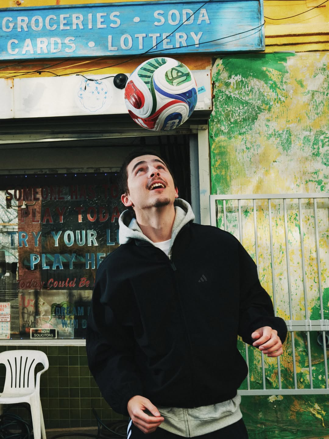 Young man in a black jacket looking up as a colorful soccer ball floats above his head outside a corner grocery store.
