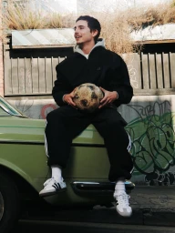Young man in a black Adidas tracksuit and sneakers sits on the hood of an olive green vintage car holding a soccer ball.