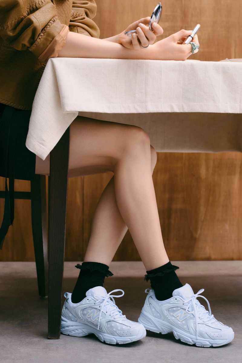 Woman seated at a table wearing white sneakers with black ruffled socks, holding a compact mirror and makeup, studio shot.