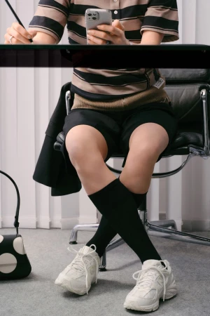 A person sits at a desk using a smartphone, dressed in a striped polo shirt, black shorts, black socks, and white sneakers with a black bag beside.