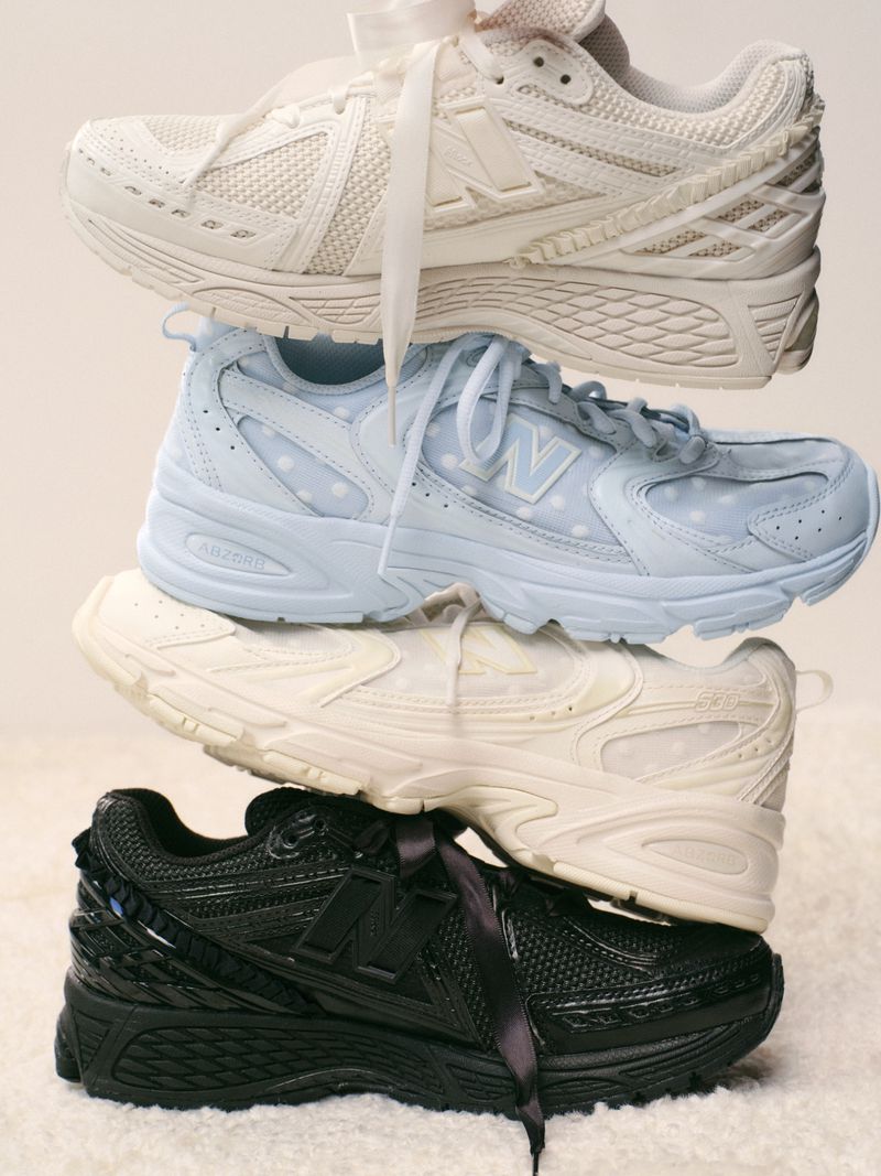 Stack of New Balance sneakers in white, light blue, cream, and black, each with a matching ribbon, on a soft, fuzzy background.