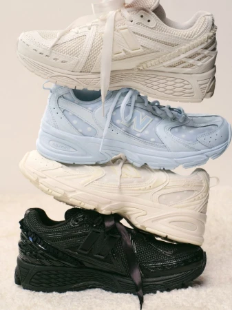 Stack of New Balance sneakers in white, light blue, cream, and black, each with a matching ribbon, on a soft, fuzzy background.