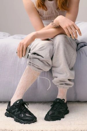 Model wearing black sneakers and grey sweatpants sitting on a bed holding a smartphone with wired headphones