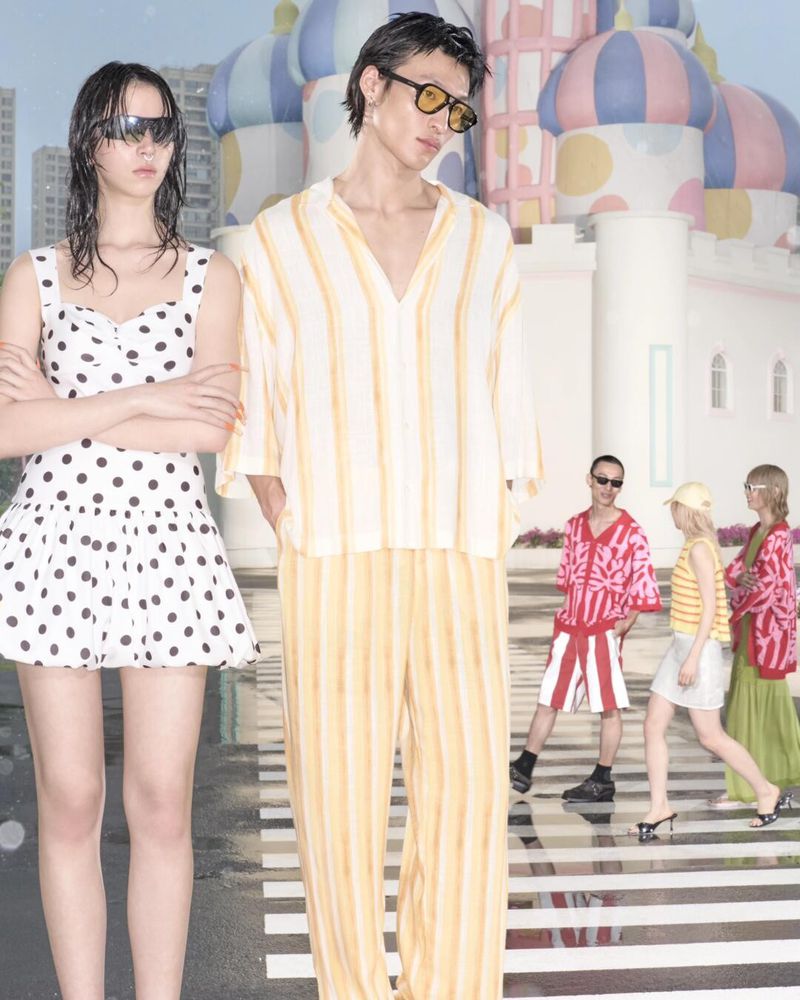 Two models posing in fashionable outfits on a city crosswalk, backed by other models and a whimsical building on a sunny day.